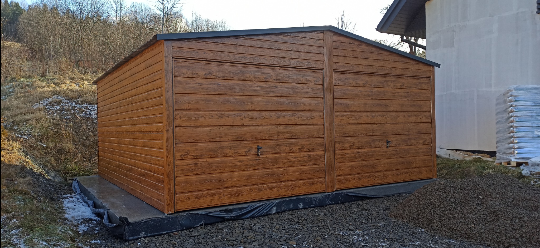 Dwustanowiskowy, brązowy garaż blaszany z imitacją drewna, umieszczony na betonowej wylewce, z widocznym otoczeniem w postaci ziemi i kamieni oraz częściowo ośnieżonego terenu.