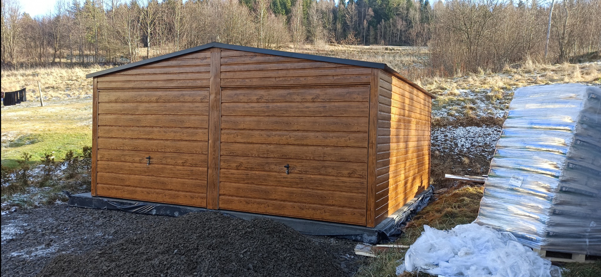 Gotowy garaż blaszany z dwoma bramami o imitacji drewna, posadowiony na fundamencie z czarnej folii, z widocznym stosem materiałów budowlanych w folii obok.