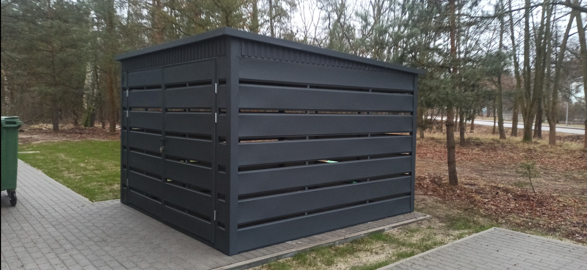 Nowoczesna, grafitowa wiata śmietnikowa z poziomymi panelami, osadzona na kostce brukowej, z widocznym pojemnikiem na śmieci po lewej stronie i drzewami w tle.