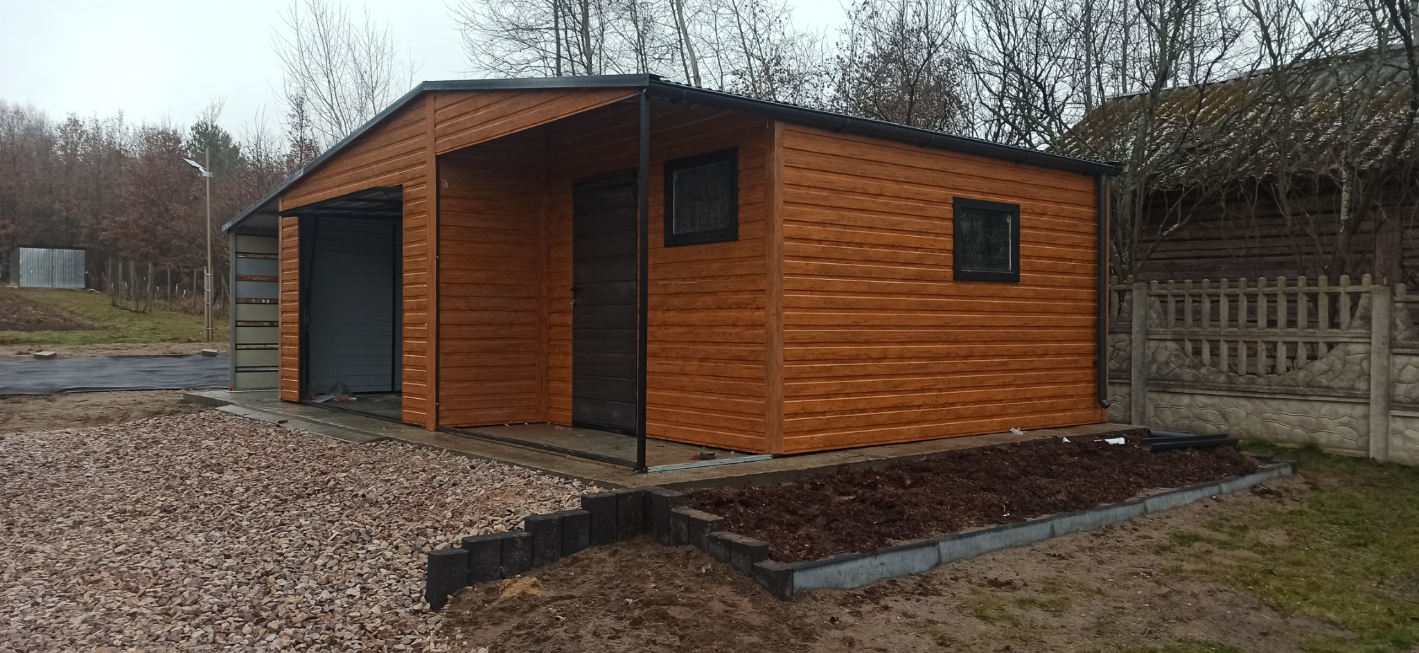 Zewnętrzny widok nowo postawionego, drewnianego garażu z bramą segmentową i drzwiami wejściowymi, otoczonego żwirem i rabatą z ziemią, na tle ogrodzenia i drzew bez liści.