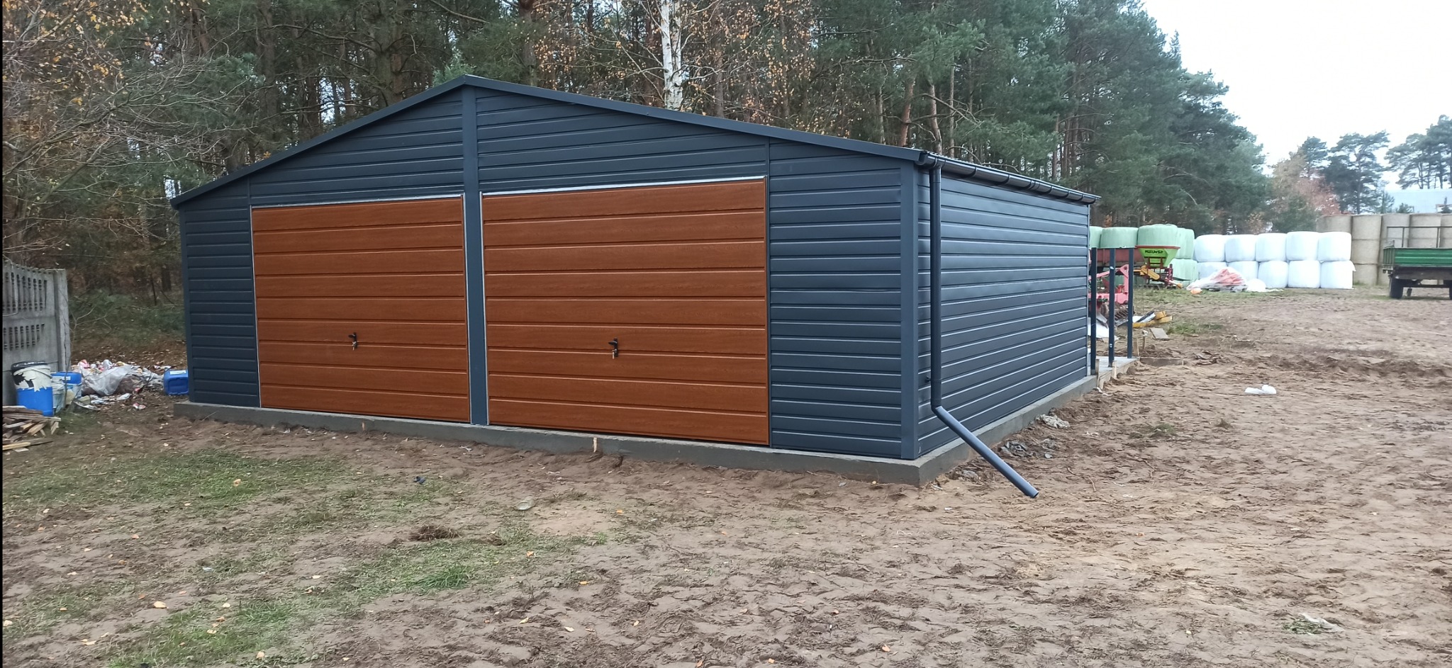 Dwustanowiskowy garaż blaszany z brązowymi bramami segmentowymi o strukturze drewna, widoczny na tle jesiennego krajobrazu.