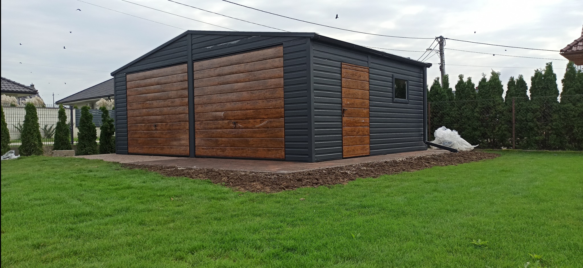 Dwustanowiskowy garaż blaszany z dwoma uchylnymi bramami o drewnopodobnej fakturze i drzwiami serwisowymi, szara blacha trapezowa, widoczne rynny i okno, otoczenie: trawnik i częściowo przygotowany...