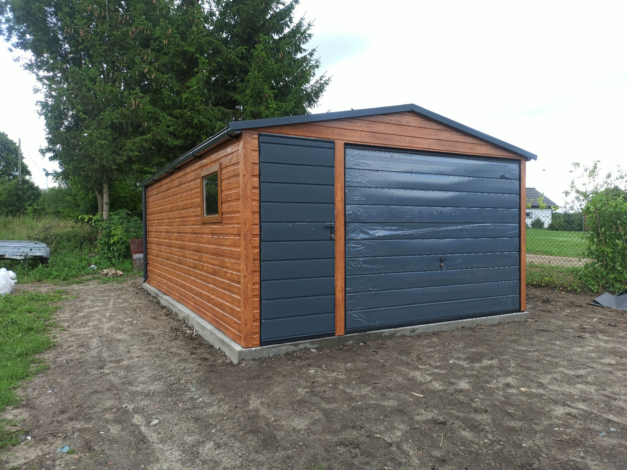 Drewniany garaż blaszany z szarą bramą segmentową i oknem, otoczony zielenią. Widoczne ślady po deszczu na bramie. Ujęcie z boku, na ziemi.