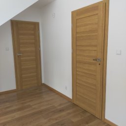 VOK - Dwa drewniane drzwi wewnętrzne z poziomymi panelami i srebrnymi klamkami, zamontowane w nowym, białym pokoju z podłogą z paneli w odcieniach brązu.