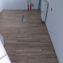 VOK - Ułożona podłoga z płytek imitujących drewno w jasnym pomieszczeniu, widoczne rury instalacyjne na ścianie i grzejnik bez maskownicy.