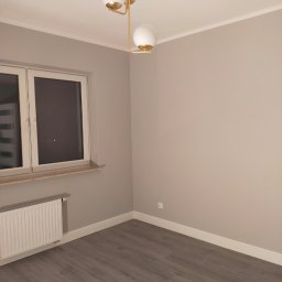 VOK - Pomalowane na szaro wnętrze pokoju z panelami podłogowymi, białym grzejnikiem i nowoczesną lampą sufitową z białymi kloszami i złotymi elementami.