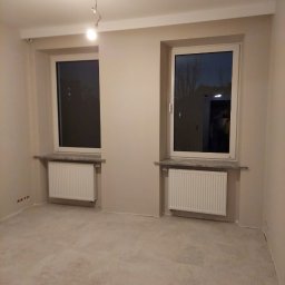 VOK - Pokój po remoncie z dwoma oknami, grzejnikami i beżowymi ścianami, widoczne ślady po gniazdkach elektrycznych w dolnej części ściany.