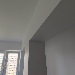 VOK - Świeżo pomalowane ściany i sufit na biało w pokoju, widoczny fragment okna z widokiem na blok mieszkalny oraz grzejnik pod oknem.