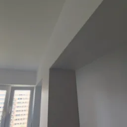 Świeżo pomalowane ściany i sufit na biało w pokoju, widoczny fragment okna z widokiem na blok mieszkalny oraz grzejnik pod oknem.