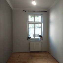 VOK - Pusty pokój po remoncie z drewnianą podłogą, świeżo pomalowanymi szarymi i białymi ścianami, białym oknem z grzejnikiem i wiszącą żarówką.