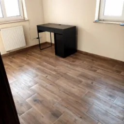 Ułożona podłoga z płytek imitujących drewno w pokoju z biurkiem i grzejnikiem pod oknem.