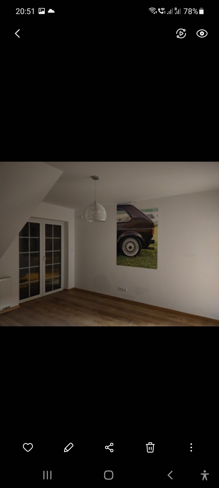 Pusty pokój z drewnianą podłogą, białymi ścianami i lampą wiszącą, na ścianie wisi obraz z brązowym samochodem.