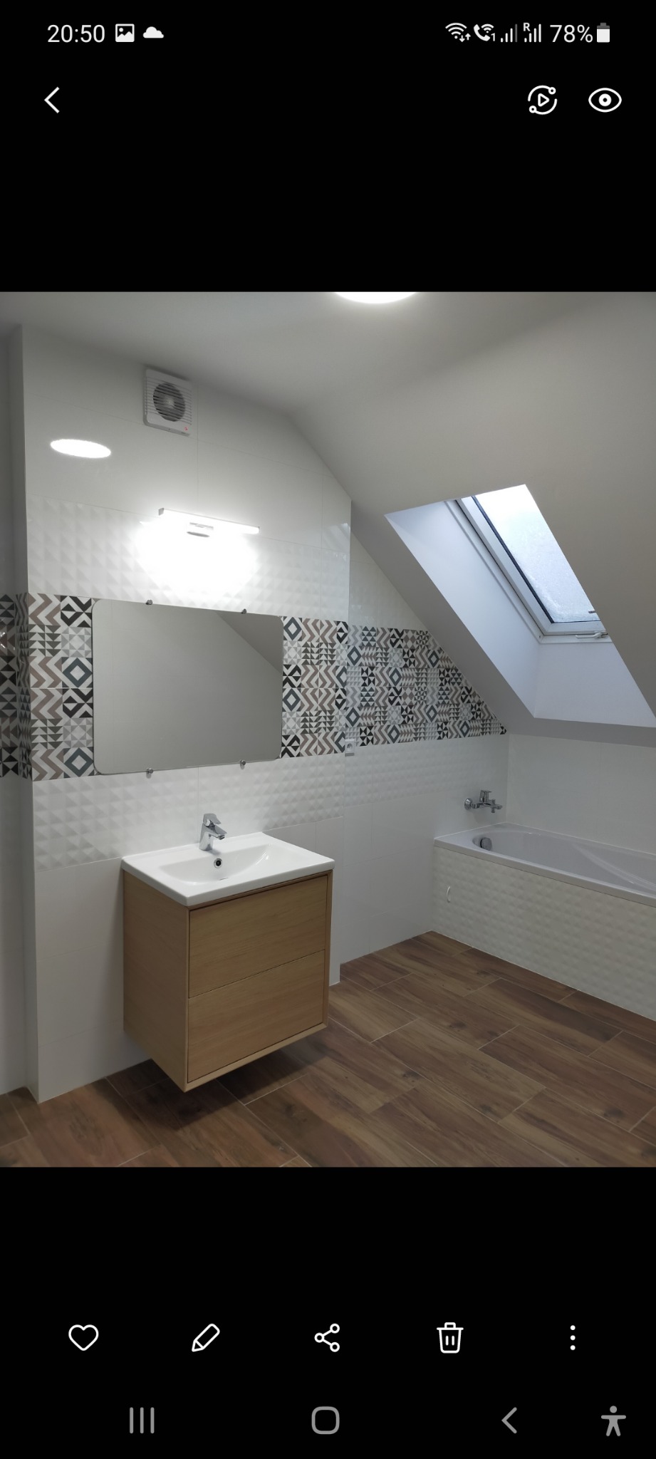 Nowoczesna łazienka na poddaszu z drewnopodobną podłogą, białą armaturą i geometrycznymi wzorami płytek na ścianach pod skosem okna dachowego.