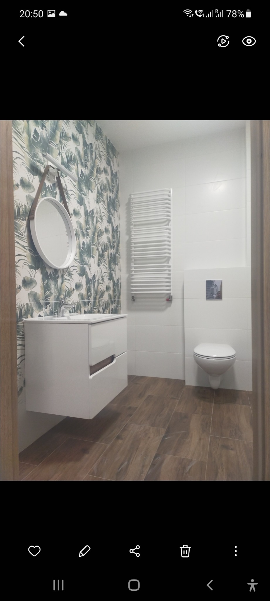 Nowoczesna łazienka z drewnopodobną podłogą, szafką pod umywalkę z białym blatem, okrągłym lustrem na pasku, grzejnikiem drabinkowym i toaletą podwieszaną na tle białych i wzorzystych płytek.