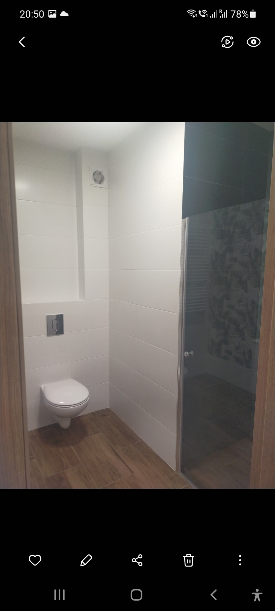 Nowoczesna łazienka z białymi, poziomymi płytkami, podwieszaną toaletą z chromowanym przyciskiem, drewnopodobną podłogą i szklaną kabiną prysznicową z ciemnymi płytkami wewnątrz.