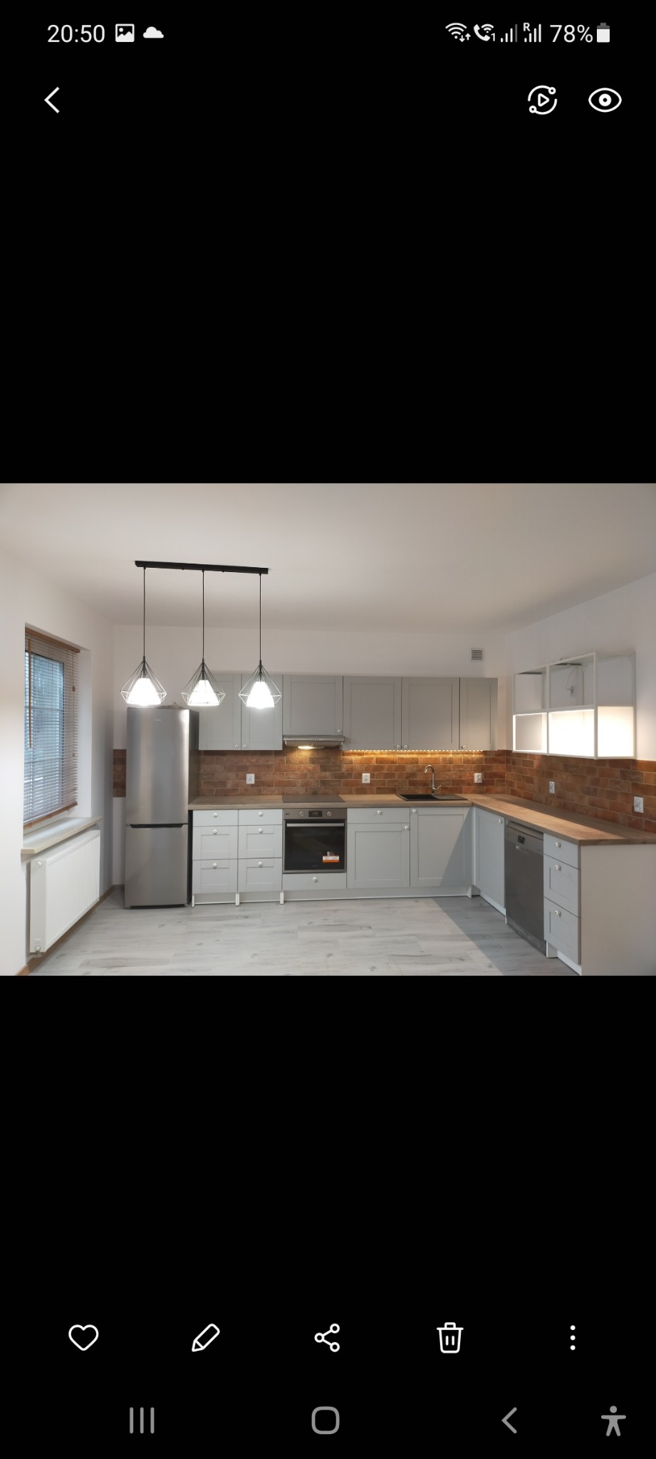 Jasna kuchnia z szarymi szafkami, ceglaną ścianą nad blatem, trzema lampami wiszącymi i drewnianymi blatami, widok na okno z żaluzjami.