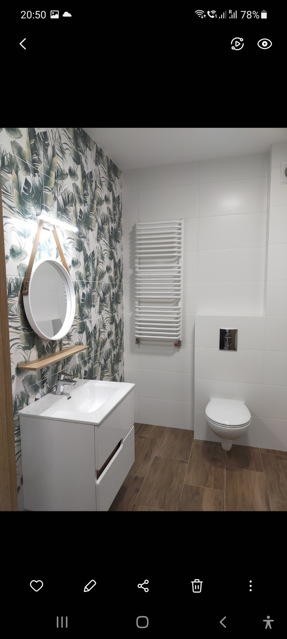 Nowoczesna łazienka z białą umywalką z szafką, okrągłym lustrem na skórzanym pasku, grzejnikiem drabinkowym, toaletą podwieszaną i płytkami z motywem roślinnym na jednej ścianie.