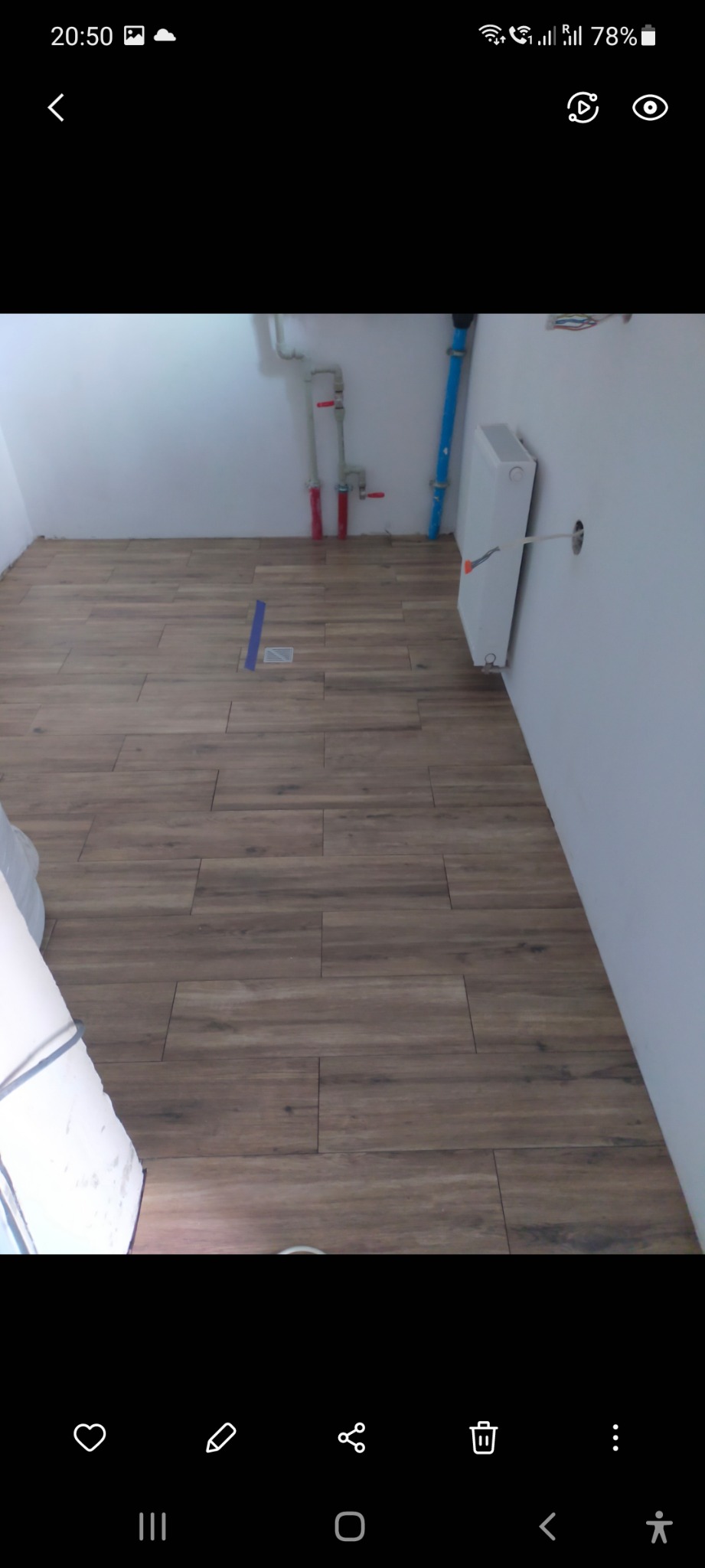 Ułożona podłoga z płytek imitujących drewno w jasnym pomieszczeniu, widoczne rury instalacyjne na ścianie i grzejnik bez maskownicy.