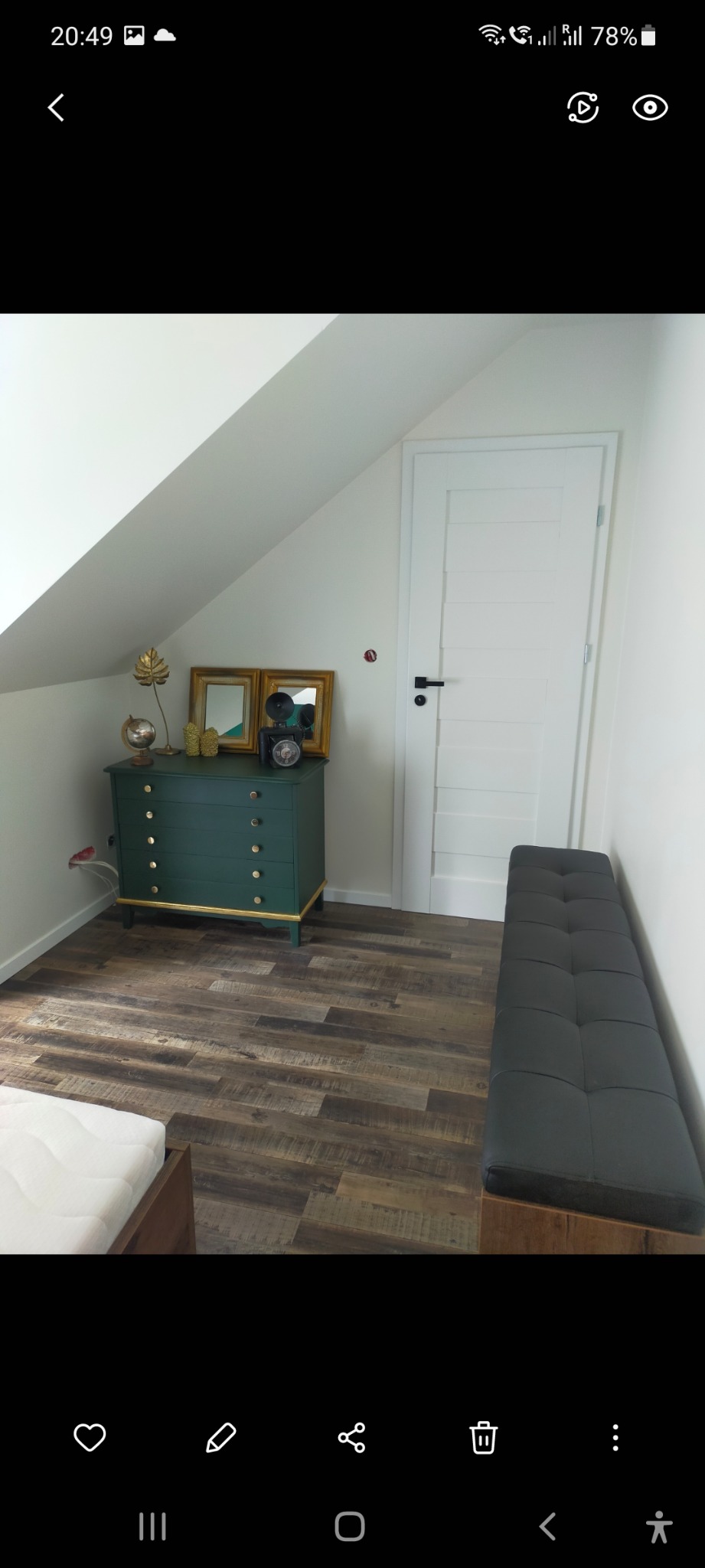 Poddasze z drewnianą podłogą, zieloną komodą z dekoracjami, białą ścianą i drzwiami oraz czarną ławką z tapicerowanym siedziskiem.