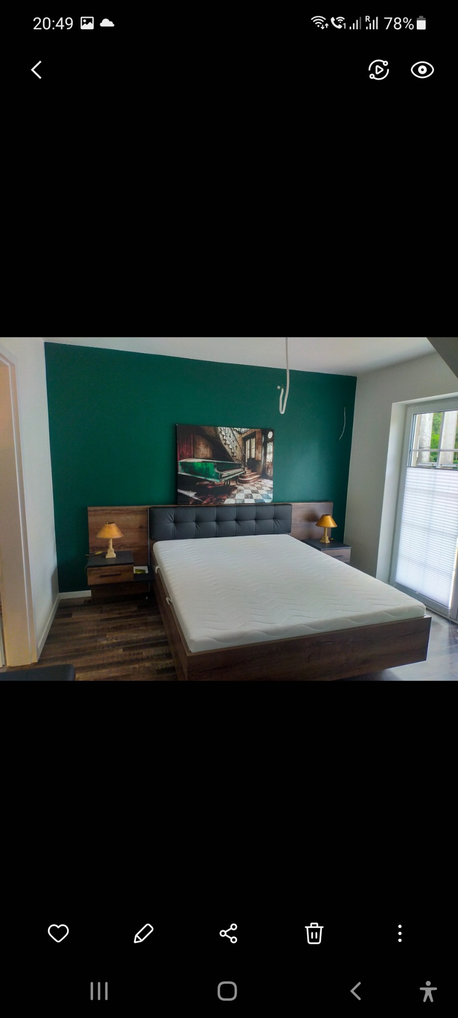 Sypialnia z łóżkiem o drewnianej ramie i tapicerowanym zagłówku, zielona ściana za łóżkiem z obrazem przedstawiającym pianino, drewniana podłoga, lampki nocne na szafkach po bokach łóżka.