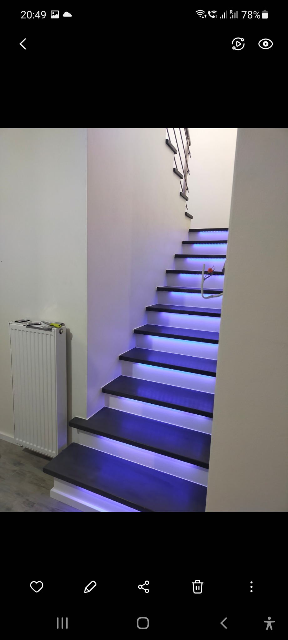 Schody z podświetleniem LED w kolorze niebieskim, czarne stopnie, białe ściany, widoczny grzejnik na ścianie po lewej stronie.