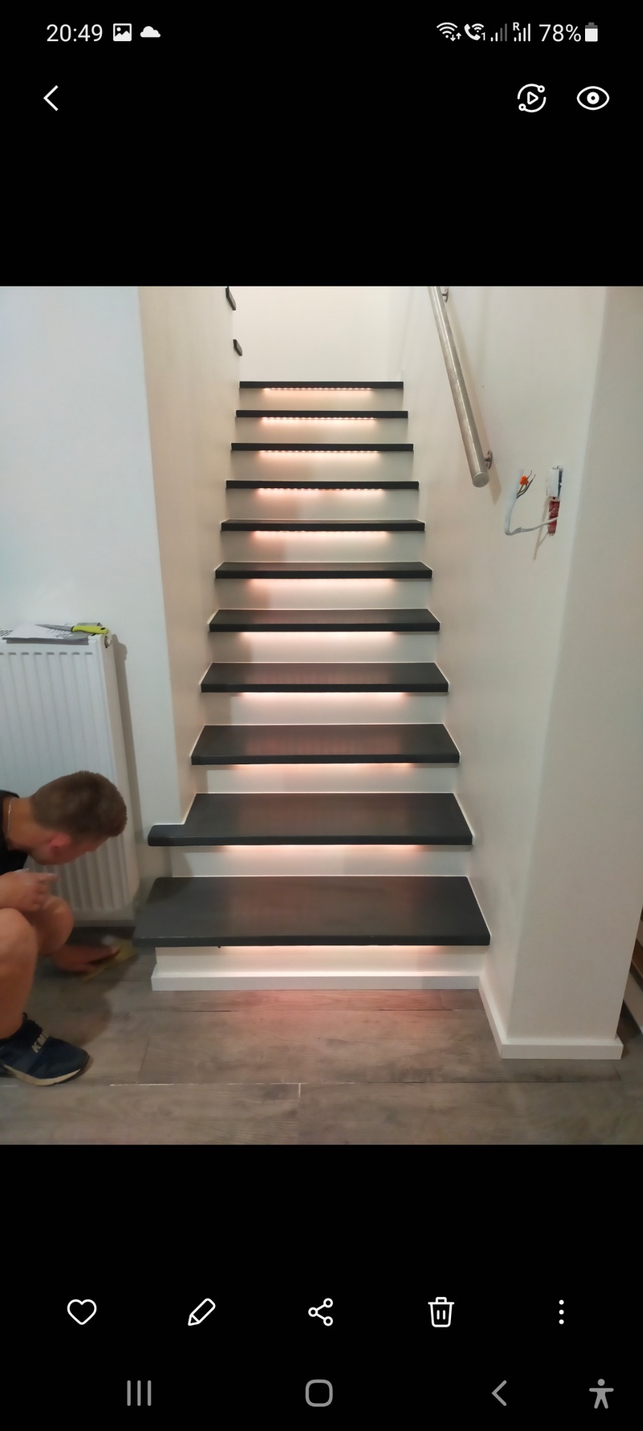 Montaż oświetlenia LED pod stopniami schodów przez pracownika, widoczne okablowanie elektryczne w ścianie i grzejnik w tle.