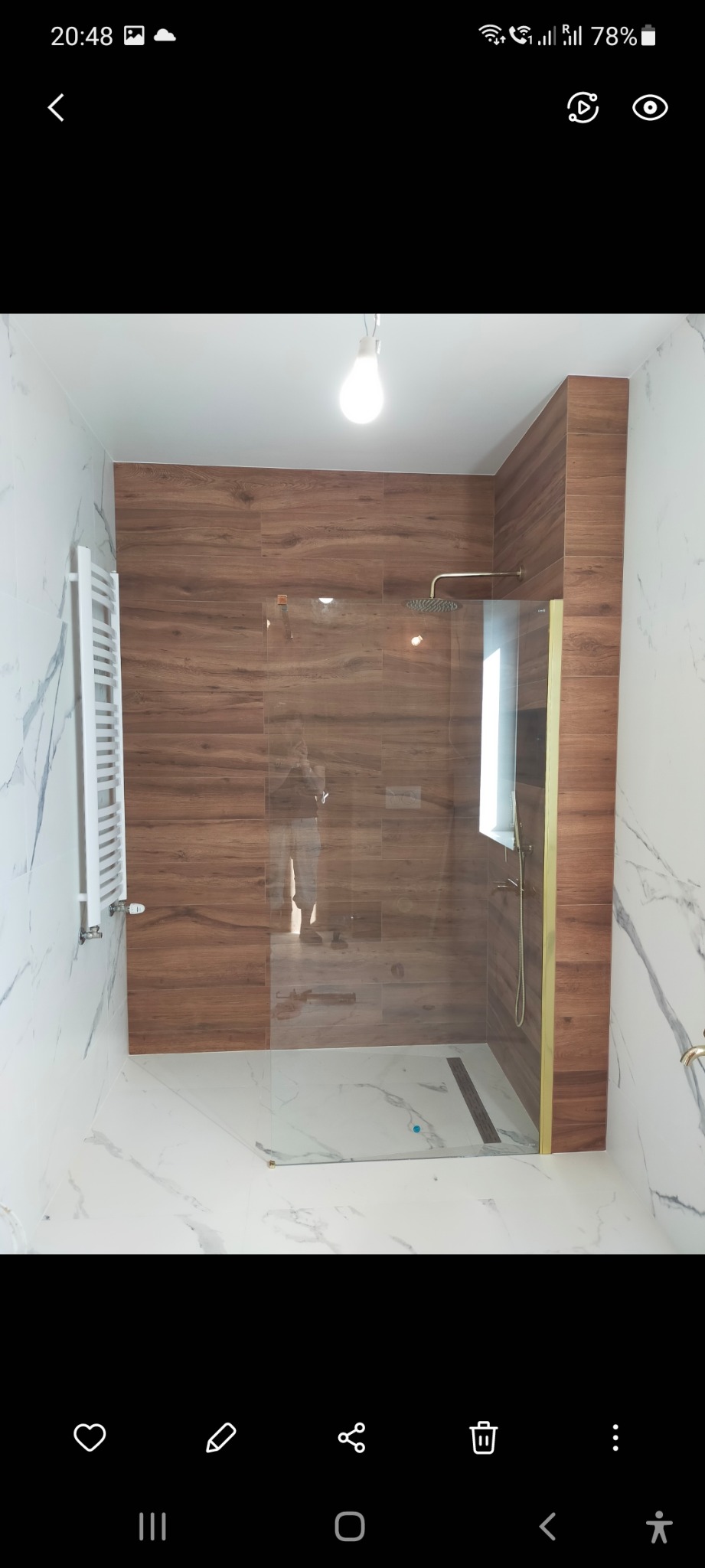 Nowoczesna kabina prysznicowa walk-in z szybą ze złotymi wykończeniami, wyłożona płytkami imitującymi drewno i marmur, z grzejnikiem drabinkowym po lewej stronie.