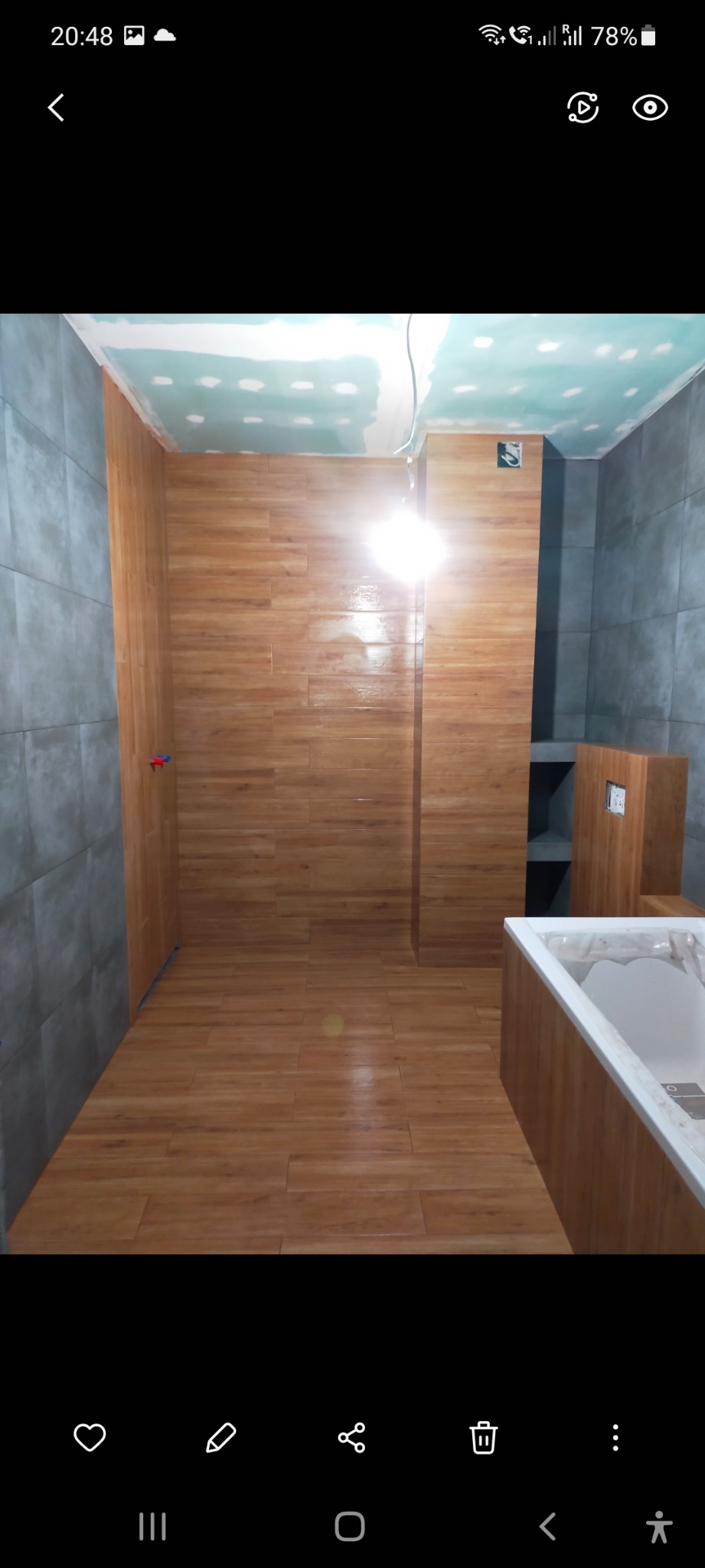 Łazienka w trakcie remontu: drewnopodobne płytki na ścianach i podłodze, surowy sufit z widocznymi punktami szpachlowania, narożna wanna częściowo obudowana, widoczne instalacje elektryczne.