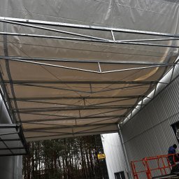 GRYCZON SPÓŁKA Z OGRANICZONĄ ODPOWIEDZIALNOŚCIĄ - Konstrukcja stalowa hali w trakcie budowy, widok od dołu na szkielet dachu z częściowo nałożonymi panelami, w tle budynek magazynowy i las.