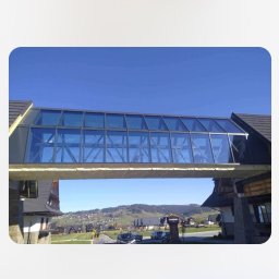 GRYCZON SPÓŁKA Z OGRANICZONĄ ODPOWIEDZIALNOŚCIĄ - Zadaszony, przeszklony łącznik między dwoma budynkami o architekturze góralskiej, na tle krajobrazu z zabudowaniami i wzgórzami.