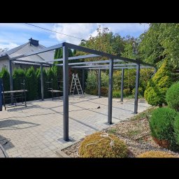 GRYCZON SPÓŁKA Z OGRANICZONĄ ODPOWIEDZIALNOŚCIĄ - Metalowa konstrukcja zadaszenia tarasu w trakcie budowy, widoczne słupy i belka poprzeczna, na tle domu i zieleni, drabina oparta o konstrukcję.