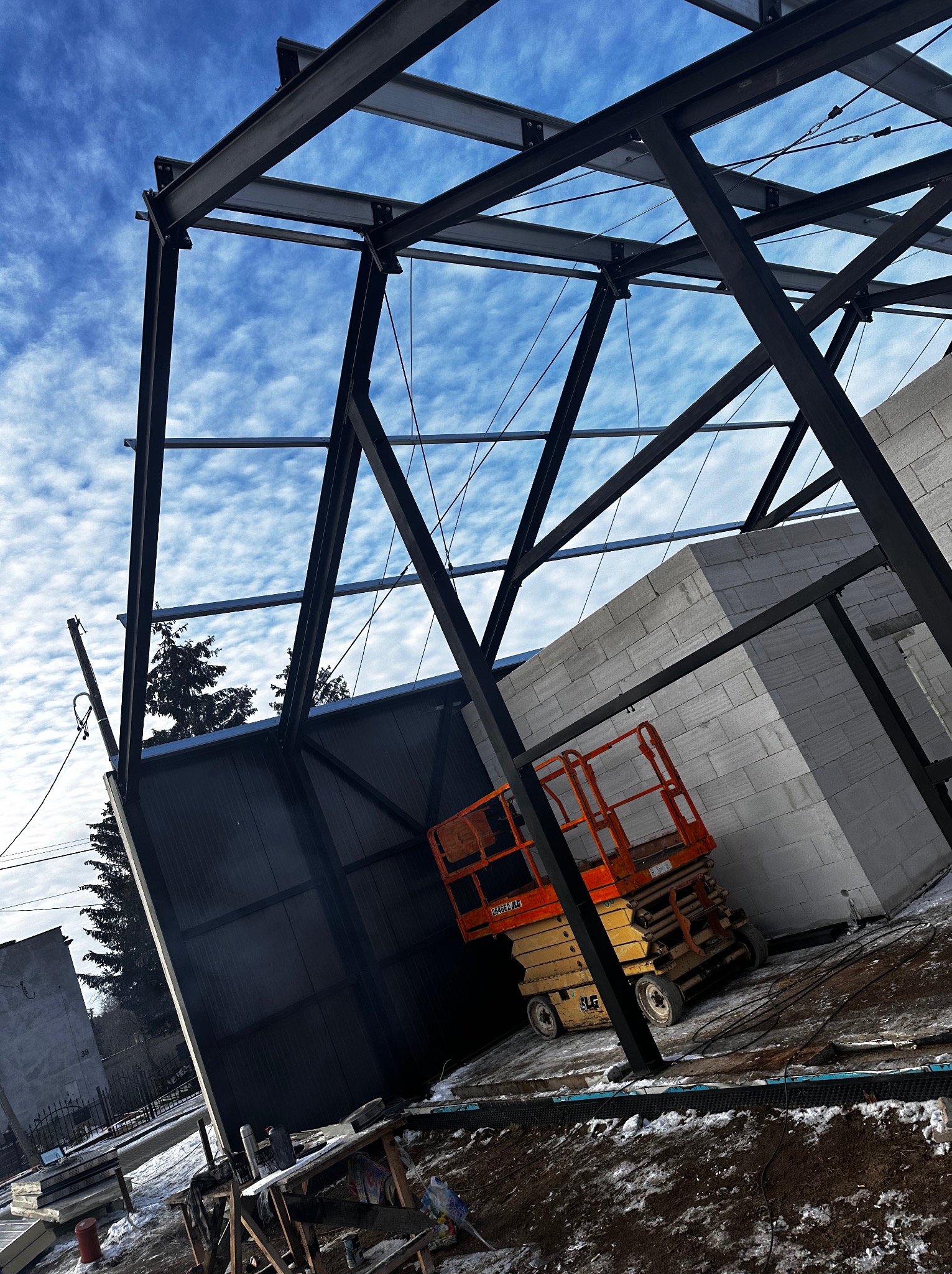 Stalowa konstrukcja hali w trakcie montażu, widoczna z dołu na tle błękitnego nieba z chmurami, obok ściana z bloczków betonowych i podnośnik koszowy.