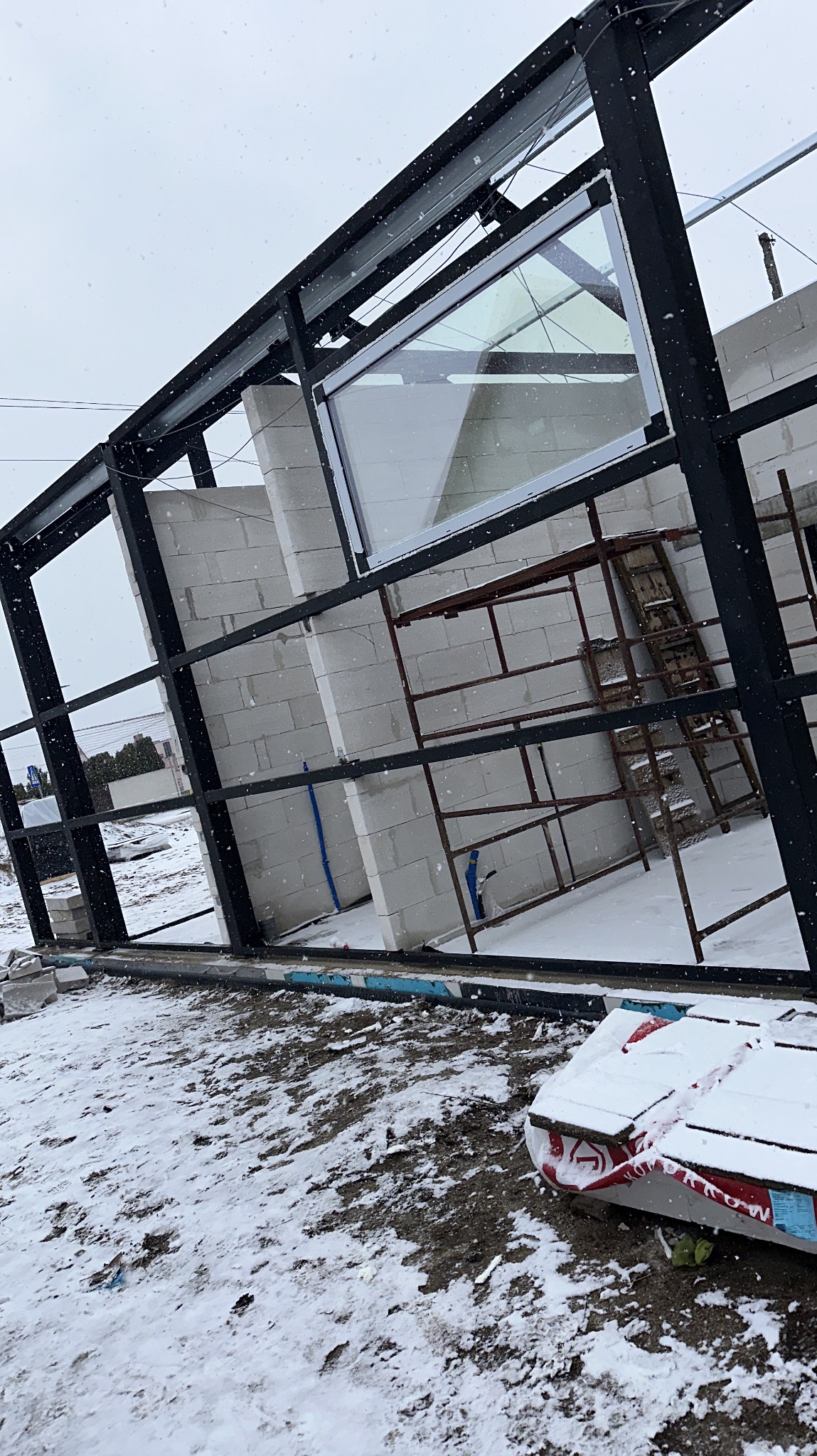 Wznoszona konstrukcja stalowa hali w Warszawie, częściowo obudowana bloczkami, w zimowej scenerii podczas opadów śniegu. Widoczne rusztowania i materiały budowlane.