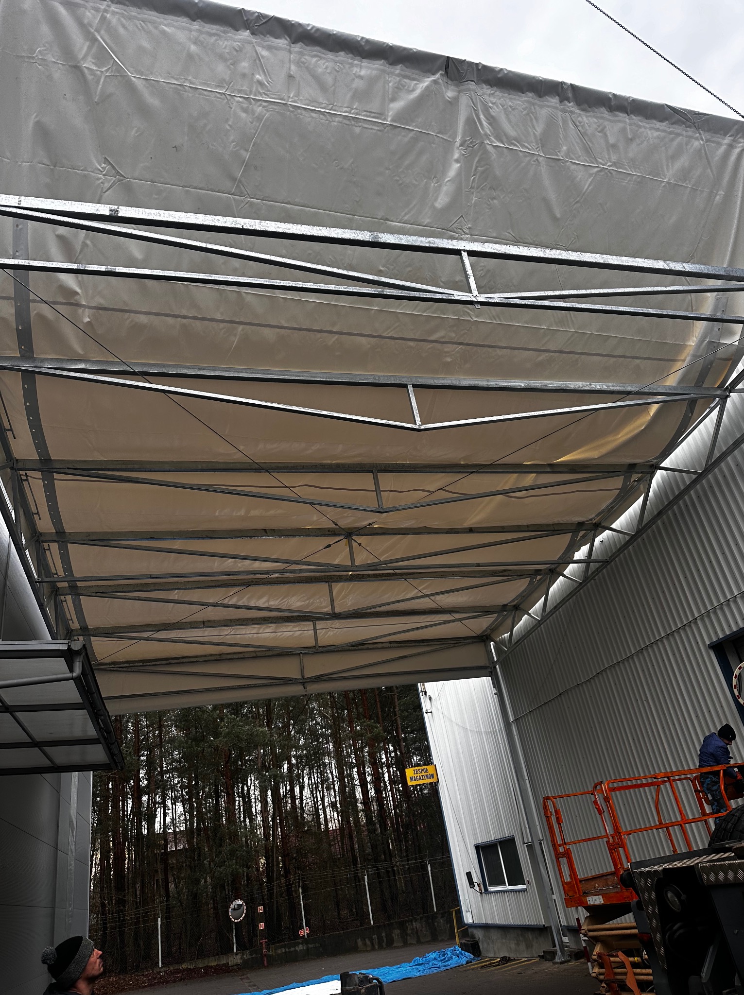 Konstrukcja stalowa hali w trakcie budowy, widok od dołu na szkielet dachu z częściowo nałożonymi panelami, w tle budynek magazynowy i las.