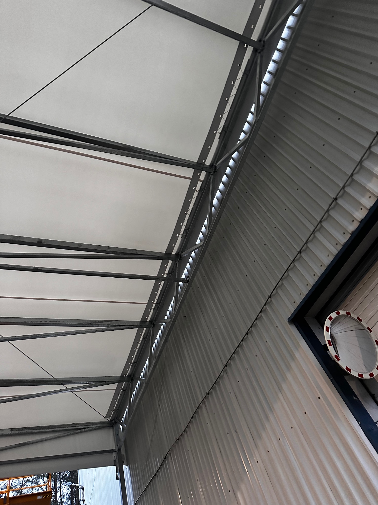 Wnętrze hali w trakcie budowy, widoczne stalowe elementy konstrukcyjne i białe płyty warstwowe, oświetlenie od góry i boczna ściana z blachy trapezowej.
