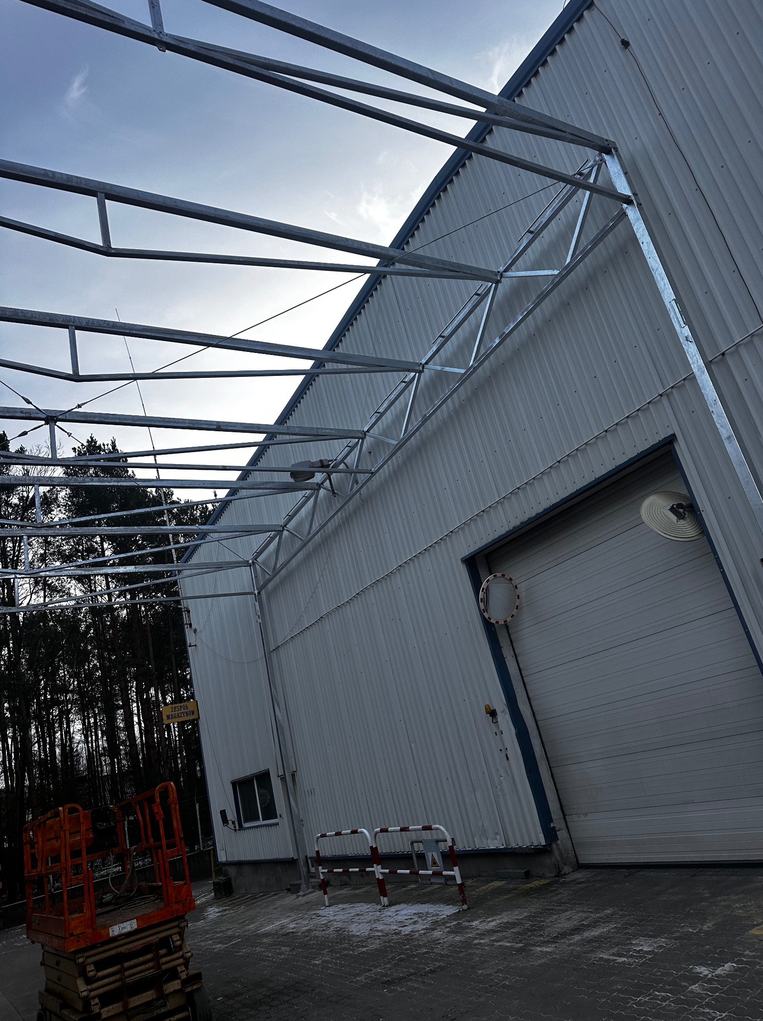 Wznoszenie konstrukcji stalowej hali magazynowej, widoczny szkielet dachu z profili stalowych, w tle ściana hali z blachy trapezowej i podnośnik koszowy.