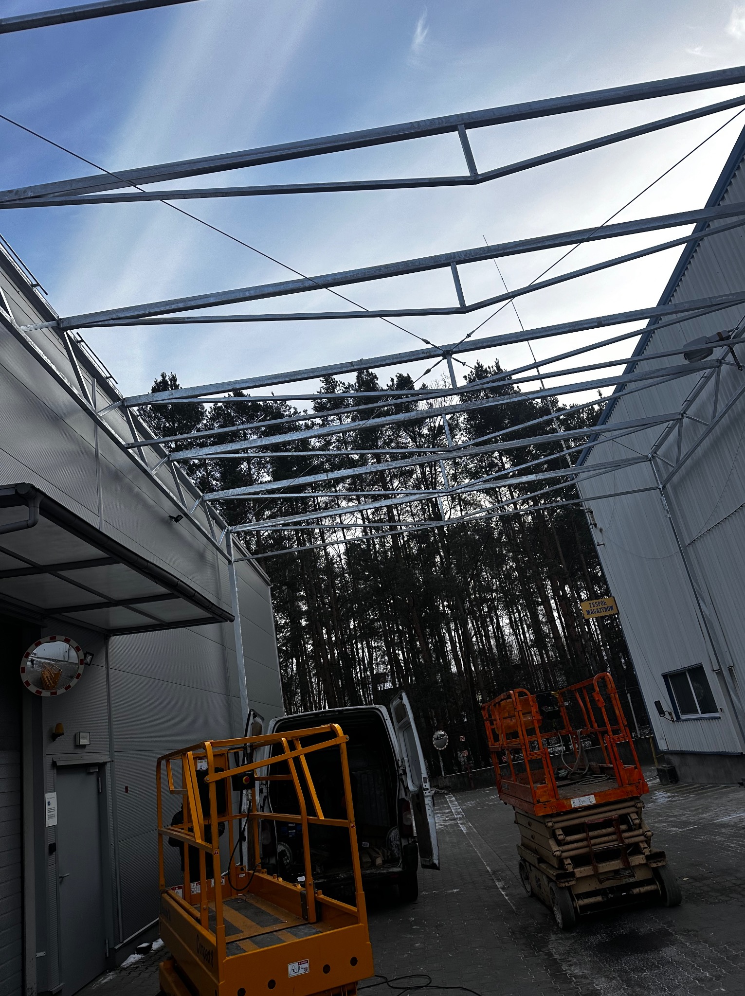 Wznoszona konstrukcja stalowa hali magazynowej, widoczne elementy szkieletu dachu, podnośniki koszowe i samochód dostawczy na zewnątrz budynku, las w tle.
