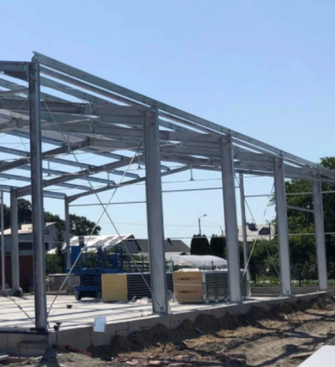 Stalowa konstrukcja hali w trakcie budowy, widoczne słupy, belki, kratownice, podłoże betonowe, materiały budowlane i podnośnik w tle.