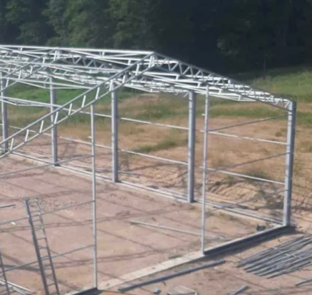 Wznoszona konstrukcja stalowa hali z widocznymi słupami, wiązarami dachowymi i stężeniami, na placu budowy z ziemistym podłożem i drabiną opartą o bok konstrukcji.