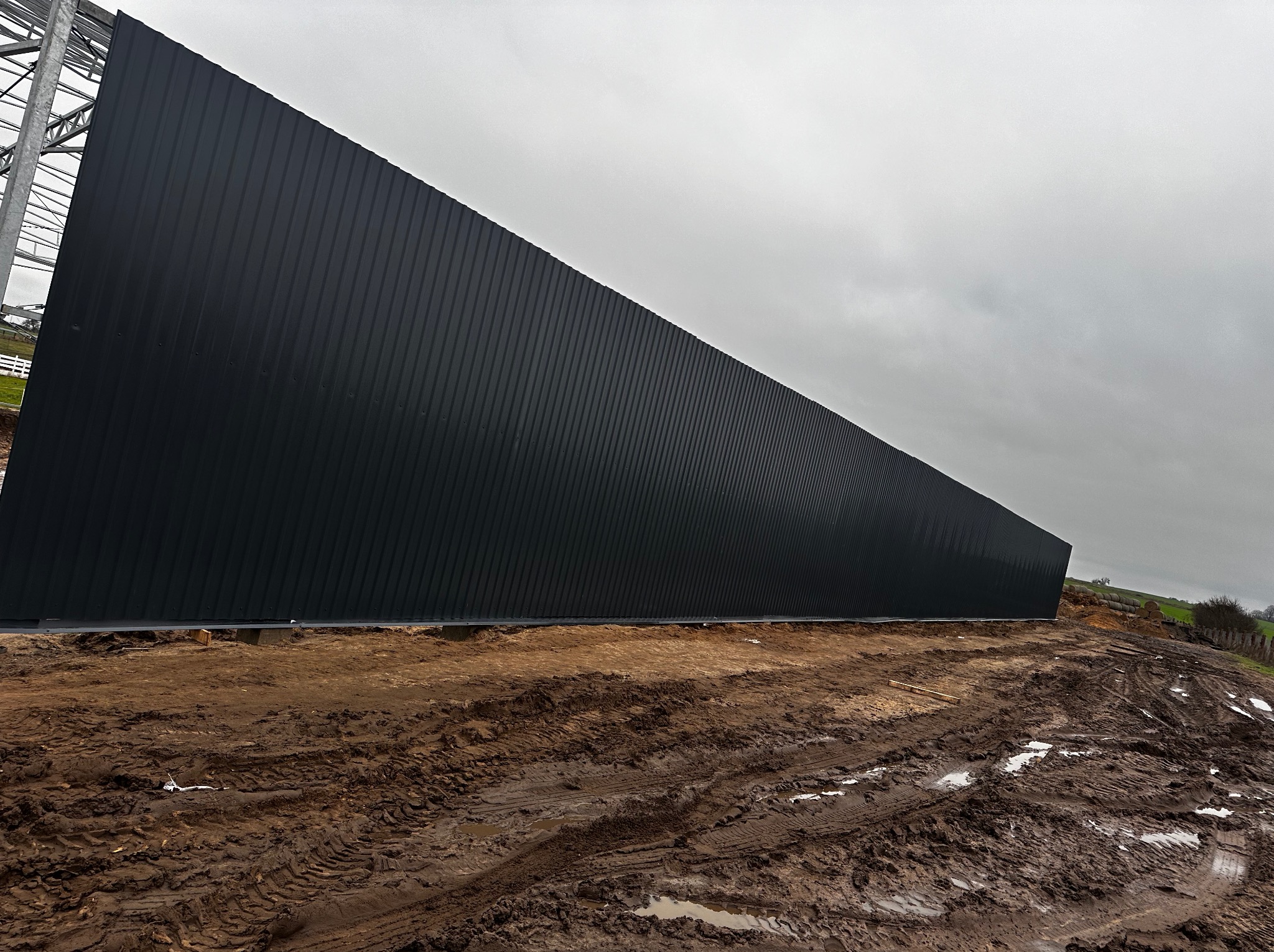 Ciemna, wysoka ściana hali z blachy trapezowej w trakcie budowy, widoczny fragment konstrukcji stalowej i błotniste podłoże po deszczu.