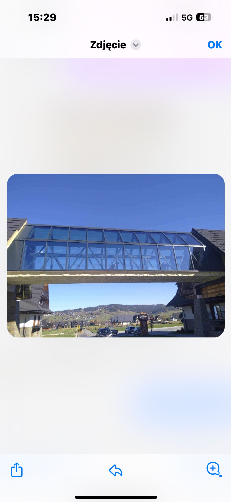 Zadaszony, przeszklony łącznik między dwoma budynkami o architekturze góralskiej, na tle krajobrazu z zabudowaniami i wzgórzami.