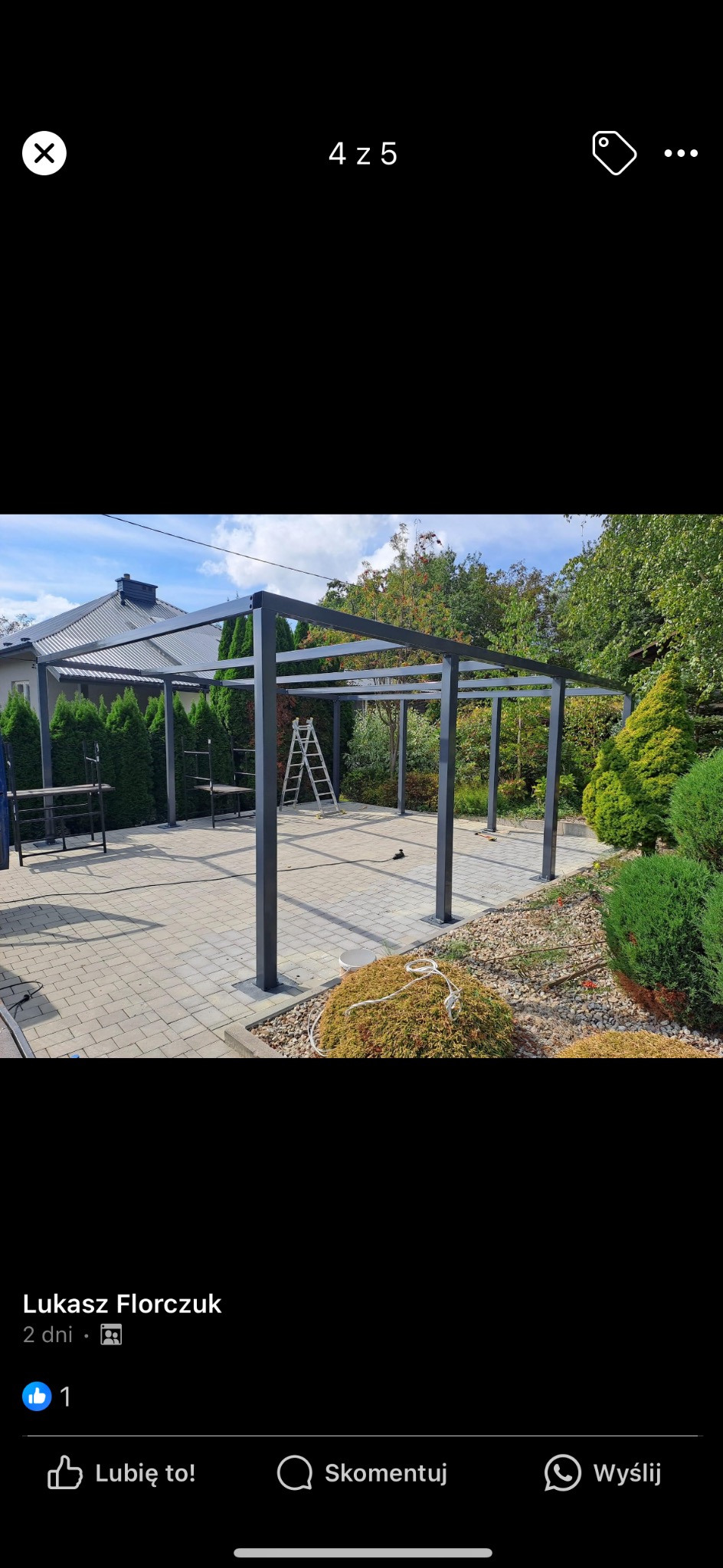 Metalowa konstrukcja zadaszenia tarasu w trakcie budowy, widoczne słupy i belka poprzeczna, na tle domu i zieleni, drabina oparta o konstrukcję.