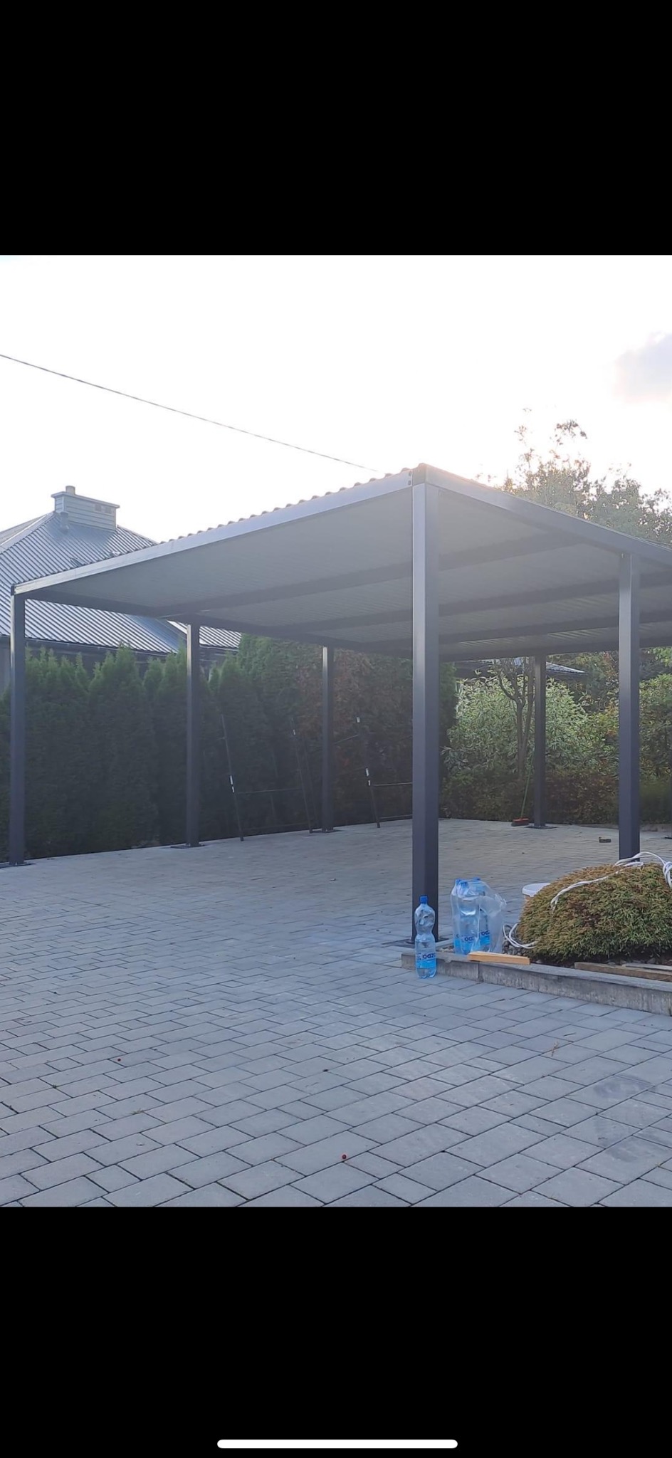 Zadaszenie tarasu lub wiaty samochodowej z konstrukcji stalowej, widoczne dwie butelki wody mineralnej i krzew ozdobny.