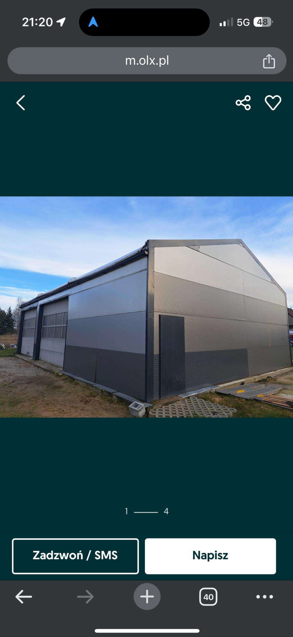 Zewnętrzny widok hali z szarej blachy falistej, z drzwiami i bramą wjazdową, na tle błękitnego nieba i drzew.