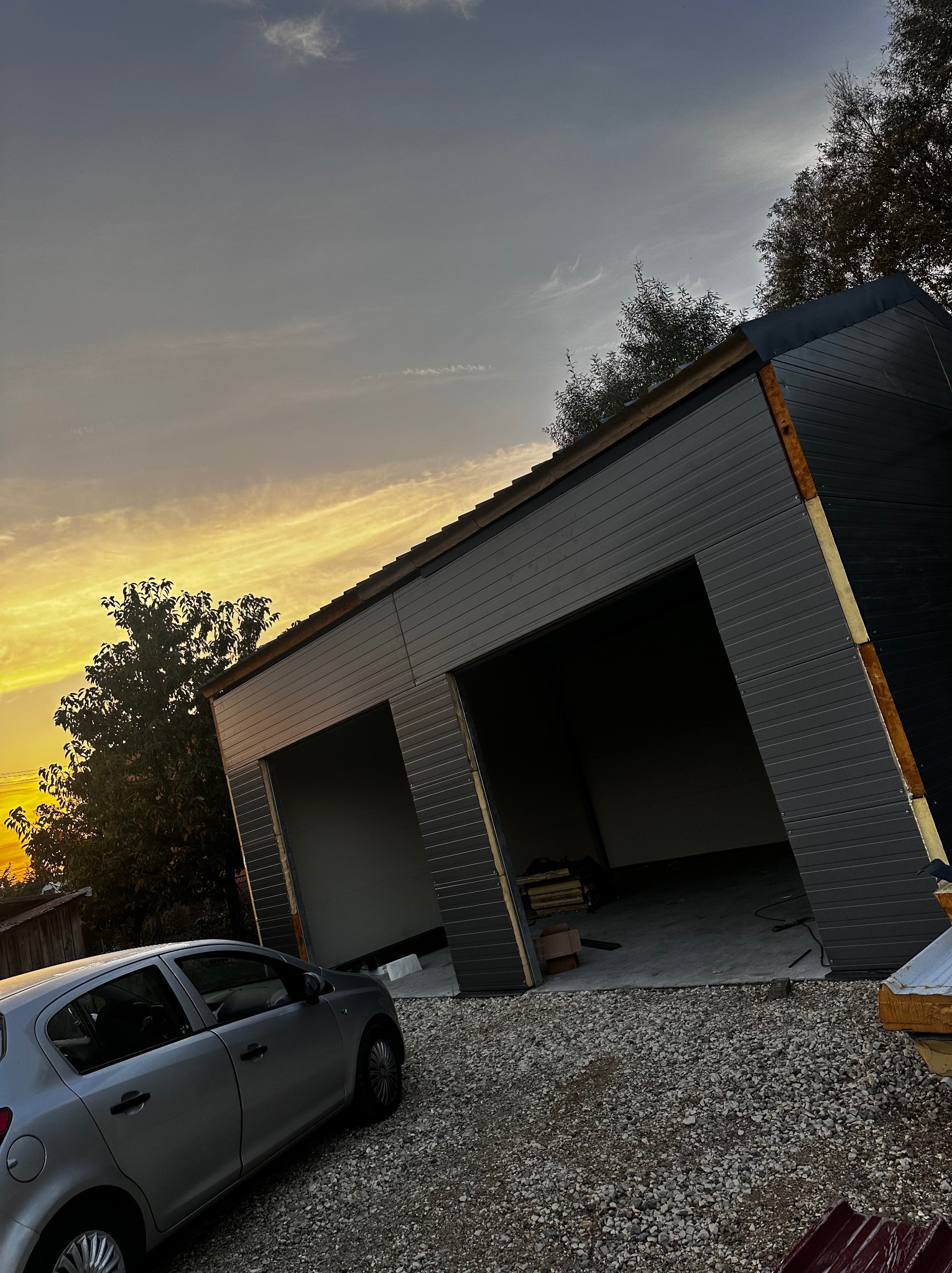 Nowoczesna hala garażowa z dwoma wjazdami, wykończona szarą blachą trapezową, widoczna na tle zachodzącego słońca i samochodu osobowego.
