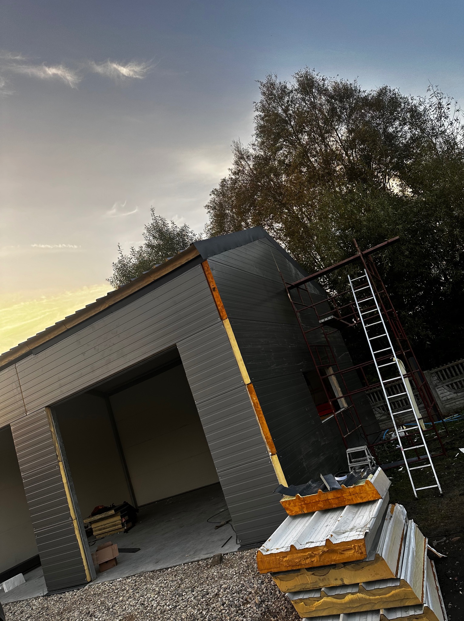 Konstrukcja hali z szarej blachy trapezowej w trakcie montażu, widoczne płyty warstwowe, rusztowanie i drabina w tle, pochmurne niebo.