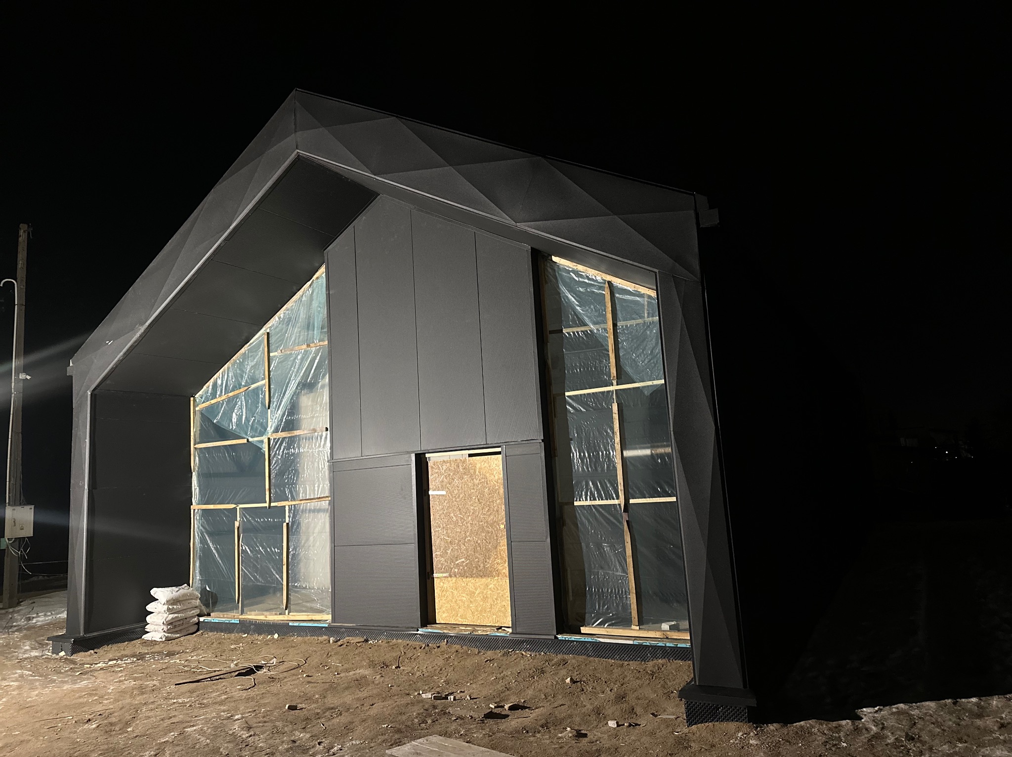 Niedokończona budowa nowoczesnej hali w nocy, z widocznymi elementami izolacji okien folią i workami z zaprawą na zewnątrz.