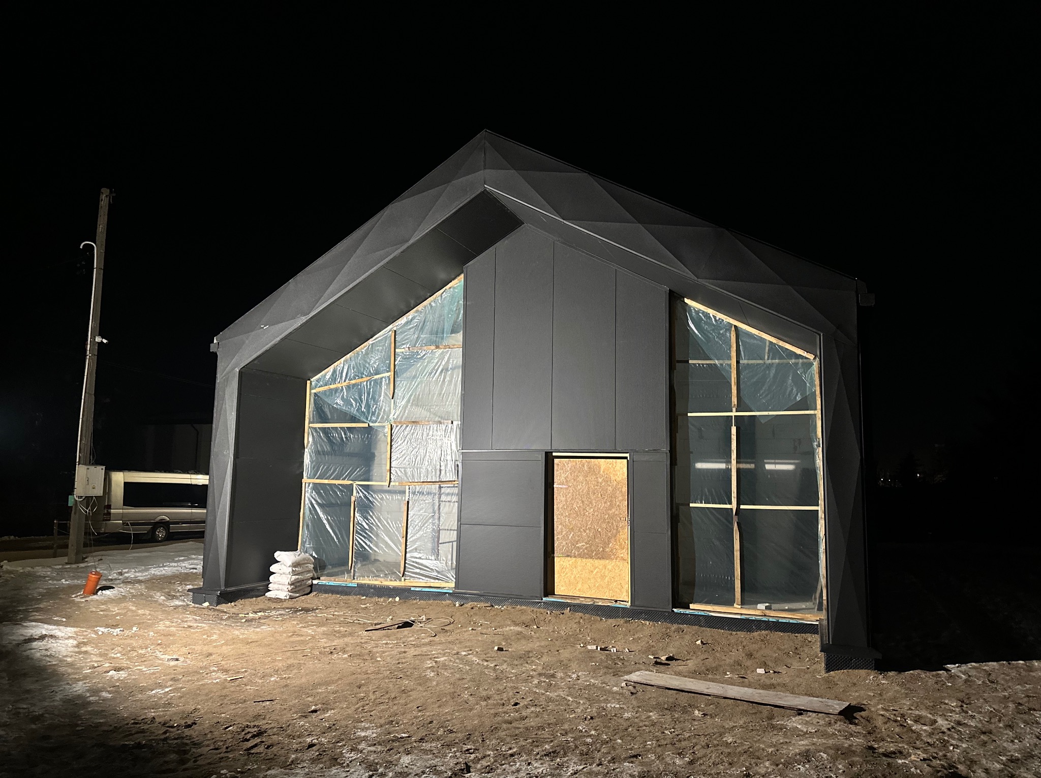 Niedokończona, nowoczesna hala z ciemnej blachy trapezowej z elementami dekoracyjnymi na dachu, okna zabezpieczone folią, nocne oświetlenie budowy.