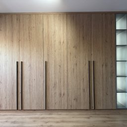 Furnilem Paweł Andrejczyn - Duża szafa wnękowa z drewnianymi frontami i czarnymi uchwytami, z otwartym regałem z podświetlanymi półkami po prawej stronie, dopasowana do drewnianej podłogi.