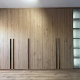 Duża szafa wnękowa z drewnianymi frontami i czarnymi uchwytami, z otwartym regałem z podświetlanymi półkami po prawej stronie, dopasowana do drewnianej podłogi.